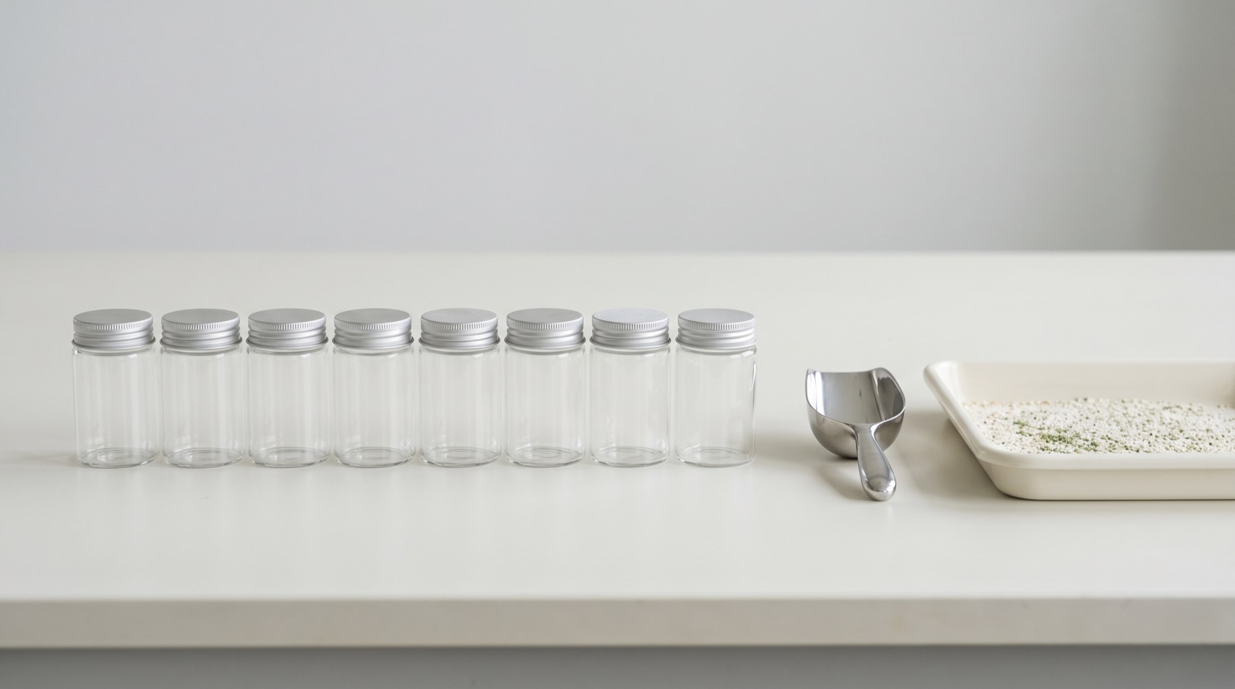 A row of small identical sample containers on a bench beside a scoop and a wide shallow tray